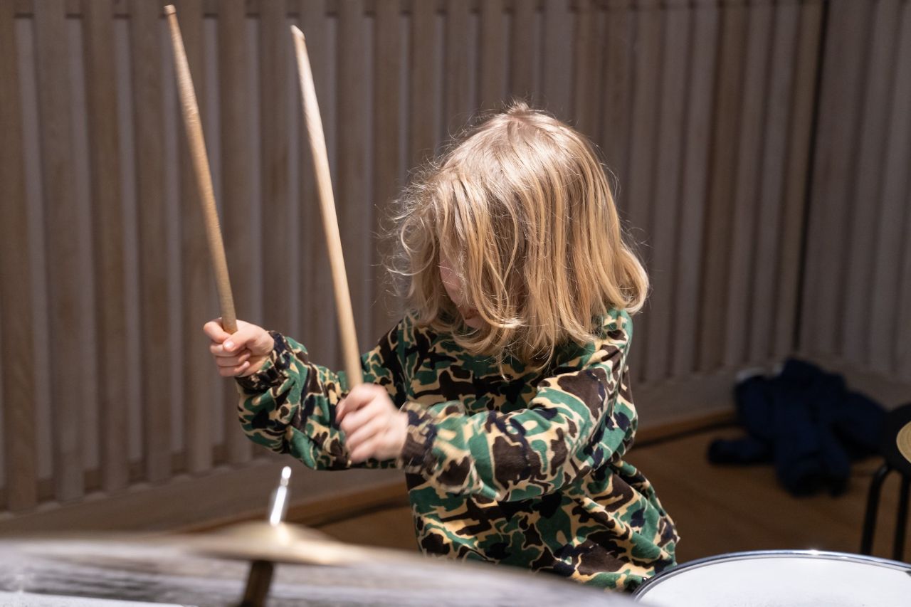 Ein Kind mit langen blonden Haaren und einem grün-braunen Pulli am Schlagzeug